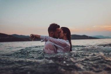 Günbatımında okyanusun dingin güzelliği arasında tutkulu bir öpücüğü paylaşan romantik genç bir çift.