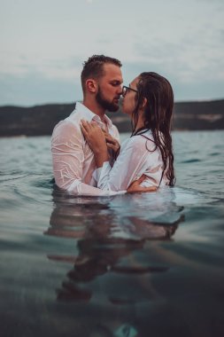 Günbatımında okyanusun dingin güzelliği arasında tutkulu bir öpücüğü paylaşan romantik genç bir çift.