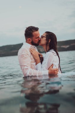 Günbatımında okyanusun dingin güzelliği arasında tutkulu bir öpücüğü paylaşan romantik genç bir çift.