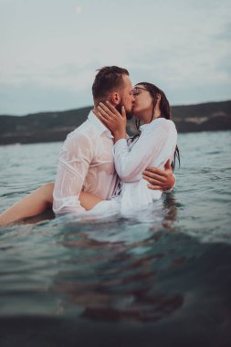 Günbatımında okyanusun dingin güzelliği arasında tutkulu bir öpücüğü paylaşan romantik genç bir çift.