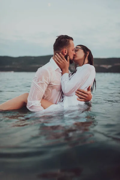 Günbatımında okyanusun dingin güzelliği arasında tutkulu bir öpücüğü paylaşan romantik genç bir çift.