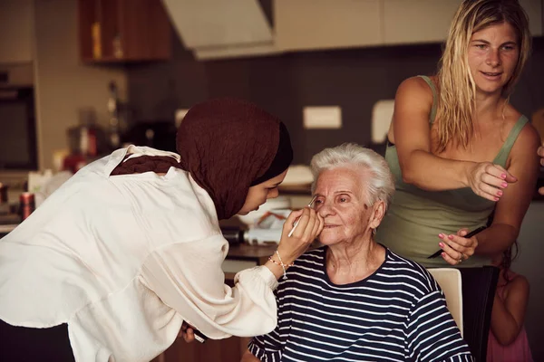 In this heartwarming real-life scene, a girl in a hijab and her sister lovingly apply makeup to their elderly grandmother, preparing her for a special family anniversary celebration, showcasing the
