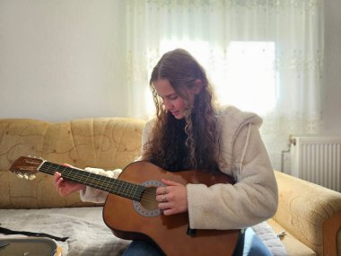 Genç, kıvırcık saçlı kız evinde tutku ve yetenekle akustik gitarını çalıyor ve bu gerçekçi fotoğrafta güzel melodiler yaratıyor..