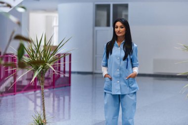 Modern siyah saçlı bir kadın doktor büyük, çağdaş bir hastanenin koridorunda kendine güvenerek duruyor, profesyonelliği ve sağlık hizmetlerine bağlılığı somutlaştırıyor.. 