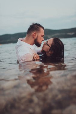 Günbatımında okyanusun dingin güzelliği arasında tutkulu bir öpücüğü paylaşan romantik genç bir çift.