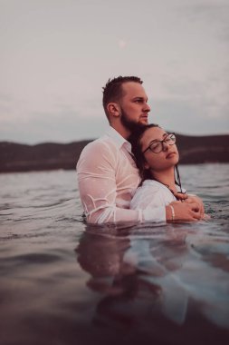 Sembolik ve duygusal olarak birbirine bağlı bir çift, sakin bir denizin arka planına nazikçe sarılır, nefes kesici bir gün batımının ılık renklerinde yıkanır.. 