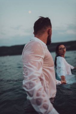 Giyinik genç bir çift, büyüleyici günbatımında denizde dururken sembolik olarak el ele tutuşarak okyanusun arka planına karşı sevgi ve birlik sahnesi yaratıyor.