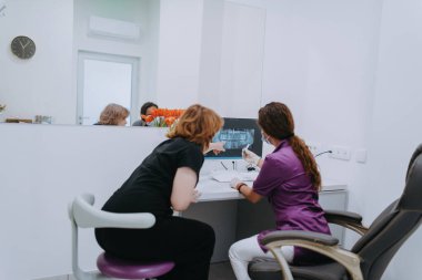 Modern bir diş muayenehanesinde, bir dişçi ve genç bir hasta yeni diş gözlüklerinin araştırılması ve bilgisayar kullanarak tedavi seçeneklerinin tartışılması gibi ortak bir konsültasyona girişirler.. 