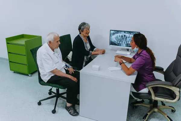 An elderly couple engages in a thoughtful discussion with their dentist about modern denture options in a contemporary dental office setting.