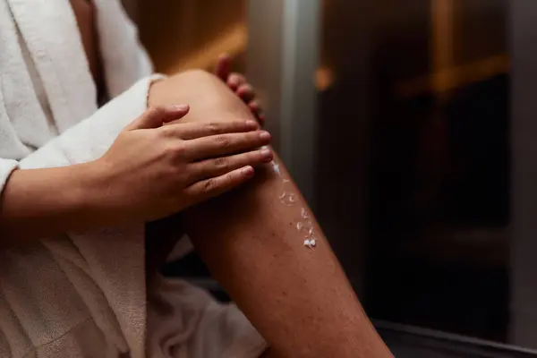A woman indulges in her self care routine, pampering her skin after a relaxing bath by applying hydrating cream to her legs, embracing the essence of beauty and well being.