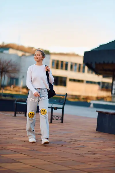 A beautiful blonde teenager returns home from school during the sunset, carrying her school backpack, in a serene and peaceful urban setting.