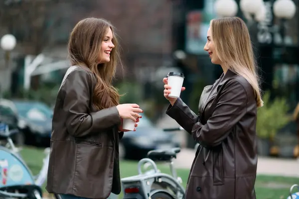 Two businesswomen take a break outside their company, enjoying a moment of relaxation as they hold cups of coffee in their hands.