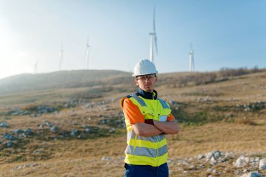 Çapraz kollu genç bir mühendis, yenilenebilir enerji projelerine güven ve adanmışlık gösteren rüzgar türbinleri alanını inceliyor.