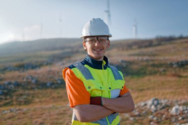 Çapraz kollu genç bir mühendis, yenilenebilir enerji projelerine güven ve adanmışlık gösteren rüzgar türbinleri alanını inceliyor.