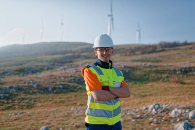 Çapraz kollu genç bir mühendis, yenilenebilir enerji projelerine güven ve adanmışlık gösteren rüzgar türbinleri alanını inceliyor.