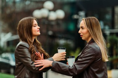 İki profesyonel kadın ofislerinin önünde duruyor, kahve içiyorlar ve molada arkadaşça sohbet ediyorlar..