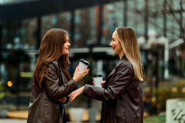 İki profesyonel kadın ofislerinin önünde duruyor, kahve içiyorlar ve molada arkadaşça sohbet ediyorlar..