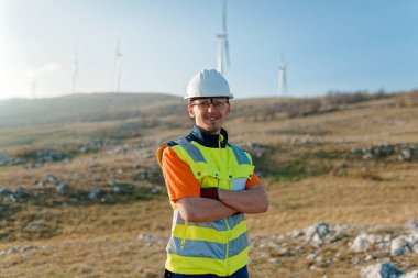 Çapraz kollu genç bir mühendis, yenilenebilir enerji projelerine güven ve adanmışlık gösteren rüzgar türbinleri alanını inceliyor.