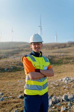 Çapraz kollu genç bir mühendis, yenilenebilir enerji projelerine güven ve adanmışlık gösteren rüzgar türbinleri alanını inceliyor.