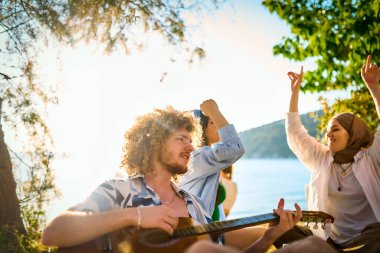 Bir grup arkadaş plajda toplanıp müzik, kahkaha ve birlikteliğin sıcaklığıyla dolu neşeli anları paylaşıyorlar. Gençler sahilde eğleniyor.