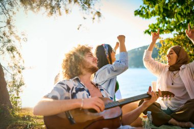 Bir grup arkadaş plajda toplanıp müzik, kahkaha ve birlikteliğin sıcaklığıyla dolu neşeli anları paylaşıyorlar. Gençler sahilde eğleniyor.