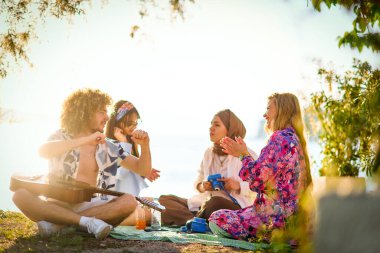 Bir grup arkadaş plajda toplanıp müzik, kahkaha ve birlikteliğin sıcaklığıyla dolu neşeli anları paylaşıyorlar. Gençler sahilde eğleniyor.