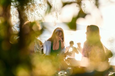 Bir grup arkadaş plajda toplanıp müzik, kahkaha ve birlikteliğin sıcaklığıyla dolu neşeli anları paylaşıyorlar. Gençler sahilde eğleniyor.