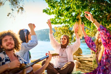 Bir grup arkadaş plajda toplanıp müzik, kahkaha ve birlikteliğin sıcaklığıyla dolu neşeli anları paylaşıyorlar. Gençler sahilde eğleniyor.