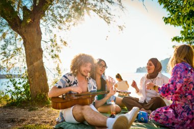 Bir grup arkadaş plajda toplanıp müzik, kahkaha ve birlikteliğin sıcaklığıyla dolu neşeli anları paylaşıyorlar. Gençler sahilde eğleniyor.
