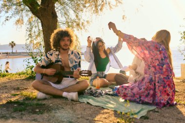 Bir grup arkadaş plajda toplanıp müzik, kahkaha ve birlikteliğin sıcaklığıyla dolu neşeli anları paylaşıyorlar. Gençler sahilde eğleniyor.