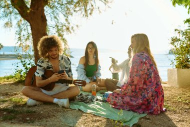 Bir grup arkadaş plajda toplanıp müzik, kahkaha ve birlikteliğin sıcaklığıyla dolu neşeli anları paylaşıyorlar. Gençler sahilde eğleniyor.