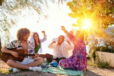 Bir grup arkadaş plajda toplanıp müzik, kahkaha ve birlikteliğin sıcaklığıyla dolu neşeli anları paylaşıyorlar. Gençler sahilde eğleniyor.