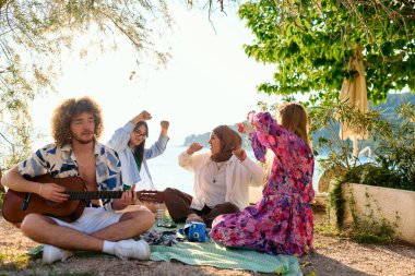 Bir grup arkadaş plajda toplanıp müzik, kahkaha ve birlikteliğin sıcaklığıyla dolu neşeli anları paylaşıyorlar. Gençler sahilde eğleniyor.