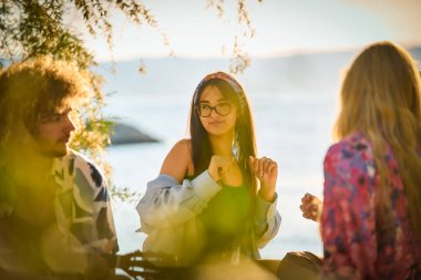 Bir grup arkadaş plajda toplanıp müzik, kahkaha ve birlikteliğin sıcaklığıyla dolu neşeli anları paylaşıyorlar. Gençler sahilde eğleniyor.