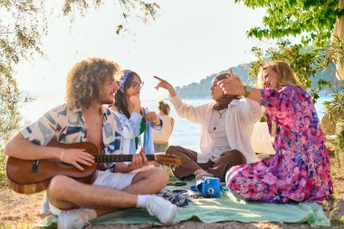 Bir grup arkadaş plajda toplanıp müzik, kahkaha ve birlikteliğin sıcaklığıyla dolu neşeli anları paylaşıyorlar. Gençler sahilde eğleniyor.