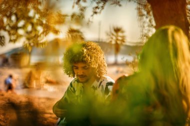 Bir grup arkadaş plajda toplanıp müzik, kahkaha ve birlikteliğin sıcaklığıyla dolu neşeli anları paylaşıyorlar. Gençler sahilde eğleniyor.