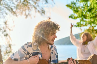 Bir grup arkadaş plajda toplanıp müzik, kahkaha ve birlikteliğin sıcaklığıyla dolu neşeli anları paylaşıyorlar. Gençler sahilde eğleniyor.