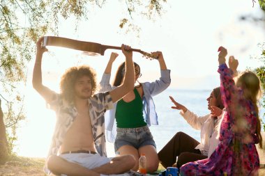 Bir grup arkadaş plajda toplanıp müzik, kahkaha ve birlikteliğin sıcaklığıyla dolu neşeli anları paylaşıyorlar. Gençler sahilde eğleniyor.