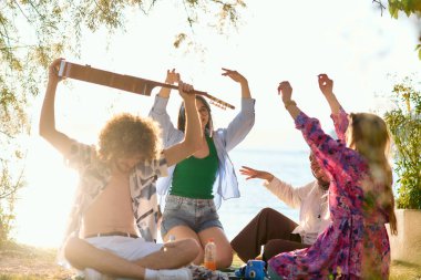 Bir grup arkadaş plajda toplanıp müzik, kahkaha ve birlikteliğin sıcaklığıyla dolu neşeli anları paylaşıyorlar. Gençler sahilde eğleniyor.