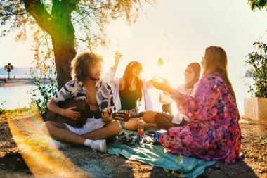 Bir grup arkadaş plajda toplanıp müzik, kahkaha ve birlikteliğin sıcaklığıyla dolu neşeli anları paylaşıyorlar. Gençler sahilde eğleniyor.