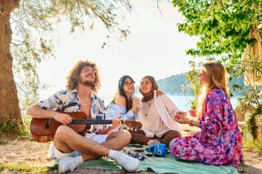 Bir grup arkadaş plajda toplanıp müzik, kahkaha ve birlikteliğin sıcaklığıyla dolu neşeli anları paylaşıyorlar. Gençler sahilde eğleniyor.