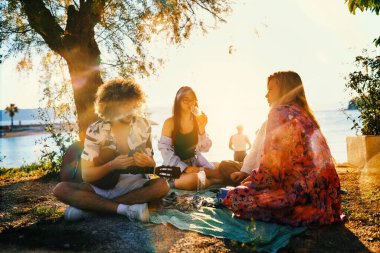 Bir grup arkadaş plajda toplanıp müzik, kahkaha ve birlikteliğin sıcaklığıyla dolu neşeli anları paylaşıyorlar. Gençler sahilde eğleniyor.