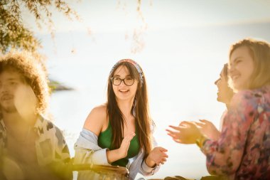 Bir grup arkadaş plajda toplanıp müzik, kahkaha ve birlikteliğin sıcaklığıyla dolu neşeli anları paylaşıyorlar. Gençler sahilde eğleniyor.