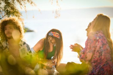 Bir grup arkadaş plajda toplanıp müzik, kahkaha ve birlikteliğin sıcaklığıyla dolu neşeli anları paylaşıyorlar. Gençler sahilde eğleniyor.