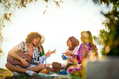 Bir grup arkadaş plajda toplanıp müzik, kahkaha ve birlikteliğin sıcaklığıyla dolu neşeli anları paylaşıyorlar. Gençler sahilde eğleniyor.