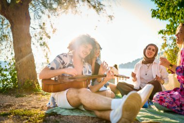 Bir grup arkadaş plajda toplanıp müzik, kahkaha ve birlikteliğin sıcaklığıyla dolu neşeli anları paylaşıyorlar. Gençler sahilde eğleniyor.