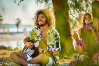 Bir grup arkadaş plajda toplanıp müzik, kahkaha ve birlikteliğin sıcaklığıyla dolu neşeli anları paylaşıyorlar. Gençler sahilde eğleniyor.