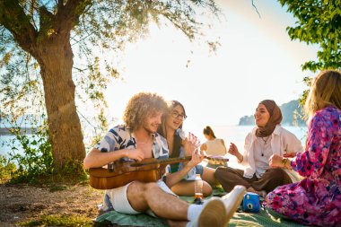 Bir grup arkadaş plajda toplanıp müzik, kahkaha ve birlikteliğin sıcaklığıyla dolu neşeli anları paylaşıyorlar. Gençler sahilde eğleniyor.