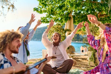Bir grup arkadaş plajda toplanıp müzik, kahkaha ve birlikteliğin sıcaklığıyla dolu neşeli anları paylaşıyorlar. Gençler sahilde eğleniyor.