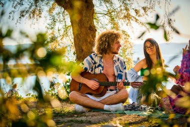 Bir grup arkadaş plajda toplanıp müzik, kahkaha ve birlikteliğin sıcaklığıyla dolu neşeli anları paylaşıyorlar. Gençler sahilde eğleniyor.